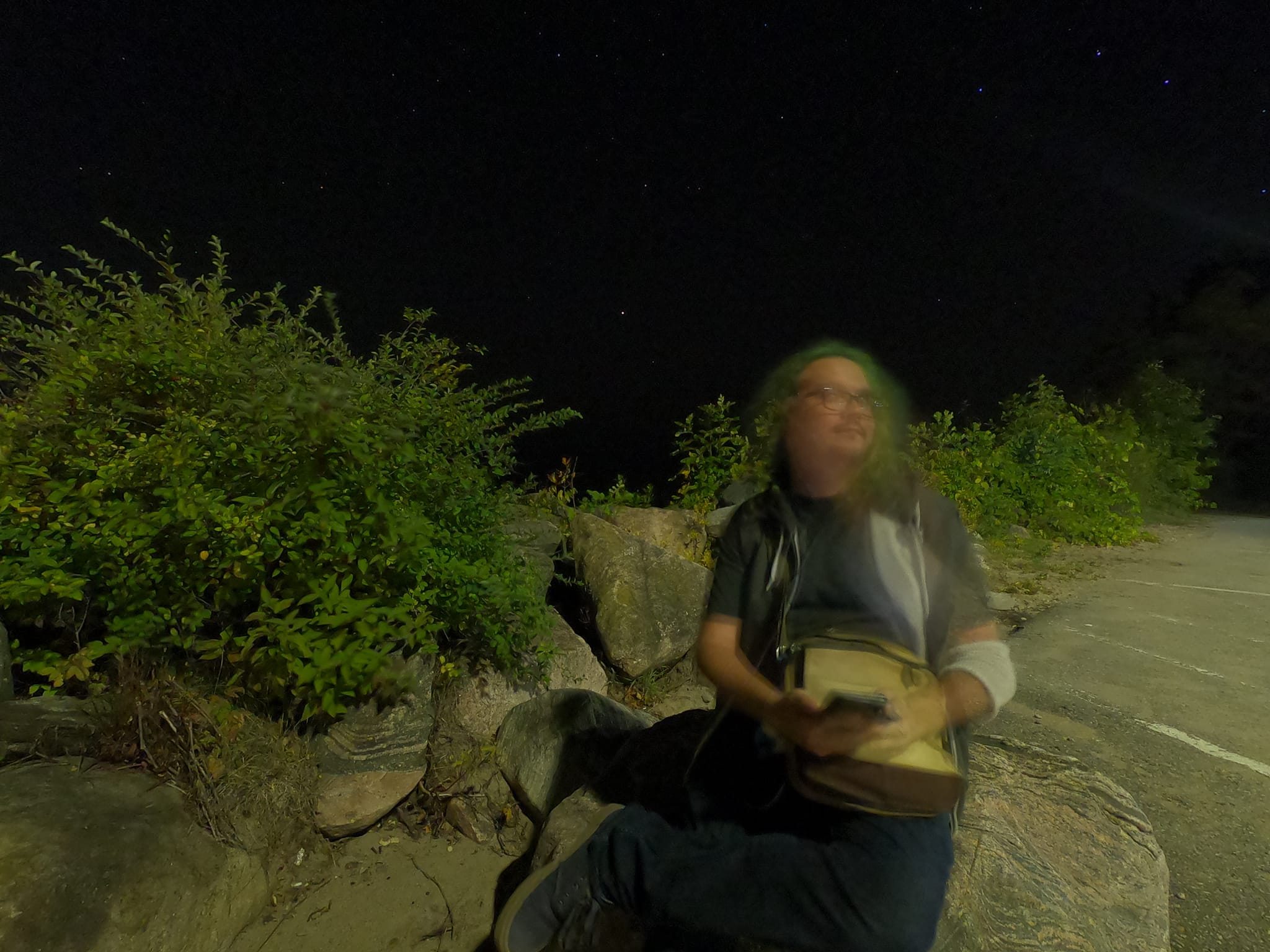 A man with long green hair and no beard sits on a rock at the Balm Beach breakwater"